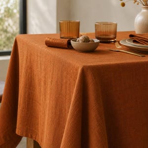 May include: A solid burnt orange tablecloth with a visible woven texture draped over a table. The table is set with two amber-colored ribbed glasses, a small ceramic bowl containing walnuts, a gold fork, and a folded matching orange napkin.