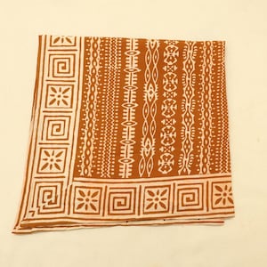 May include: A square fabric cloth featuring a burnt orange background with a white printed pattern. The design includes a border of repeating square geometric motifs and floral shapes, alongside vertical bands of intricate tribal-inspired lines.