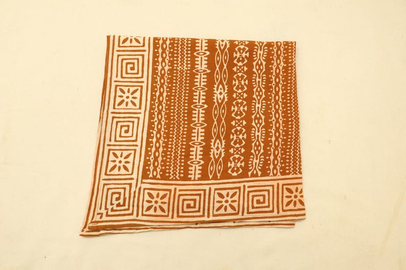 May include: A square fabric cloth featuring a burnt orange background with a white printed pattern. The design includes a border of repeating square geometric motifs and floral shapes, alongside vertical bands of intricate tribal-inspired lines.