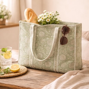 May include: A light green tote bag with a floral pattern, filled with a baguette, white flowers, and sunglasses. The bag has white handles and trim. A glass of water with lemon and mint sits on a wooden table.