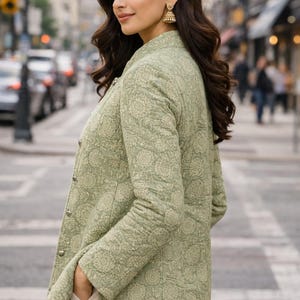 May include: A light green quilted jacket with a floral pattern. The jacket has a stand-up collar and silver buttons. The person is wearing beige pants and gold earrings. The background is a city street with cars and buildings.