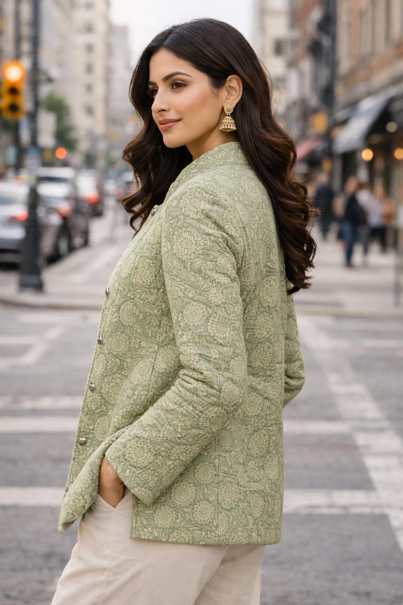May include: A light green quilted jacket with a floral pattern. The jacket has a stand-up collar and silver buttons. The person is wearing beige pants and gold earrings. The background is a city street with cars and buildings.
