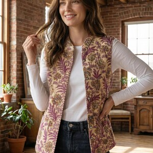 Op de afbeelding: Een gewatteerde vest met een bloemenpatroon in tinten paars, goud en crème. De vest is dichtgeknoopt en wordt gedragen over een wit shirt met lange mouwen en een donkerblauwe jeans. De vest heeft een opstaande kraag.