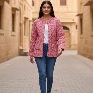 May include: A pink floral quilted jacket with a mandarin collar, worn over a white top and blue jeans. The jacket features a repeating pattern of pink flowers and green leaves. The model is wearing gold sandals.