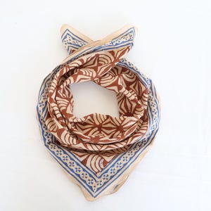 May include: A square scarf featuring a repeating brown fan-like pattern on a light beige background. The scarf is bordered by a blue and beige geometric design consisting of small cross shapes and thin lines, creating a distinct frame around the centre.