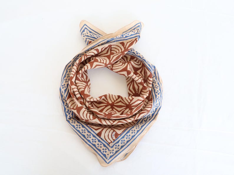 May include: A square scarf featuring a repeating brown fan-like pattern on a light beige background. The scarf is bordered by a blue and beige geometric design consisting of small cross shapes and thin lines, creating a distinct frame around the centre.