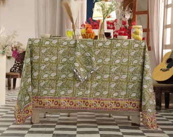 May include: A green tablecloth with a white floral pattern covers a white table. The tablecloth has a decorative border with red and yellow accents. A guitar leans against a chair in the background, and the room has a checkered floor.