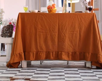 Puede incluir: Un mantel de color naranja quemado con un borde con volantes cubre una mesa puesta para una comida. La mesa está puesta con un cuenco, vasos y una cesta de naranjas. El suelo tiene un patrón de cuadros blancos y negros.