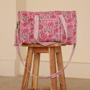 May include: A rectangular duffel bag featuring a pink and grey floral block print pattern. The bag includes two top handles and a long adjustable shoulder strap, all finished with a white and pink striped fabric trim along the edges and seams.