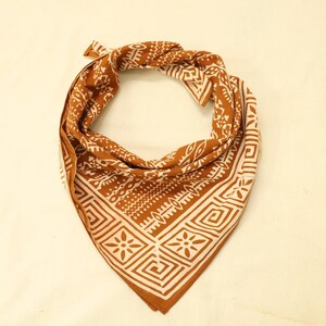 May include: A triangular bandana featuring a rust-coloured background with a white geometric pattern. The design includes repeating square motifs with central floral shapes, bordered by intricate lines and abstract symbols across the fabric.