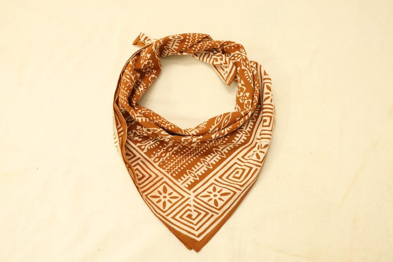 May include: A triangular bandana featuring a rust-coloured background with a white geometric pattern. The design includes repeating square motifs with central floral shapes, bordered by intricate lines and abstract symbols across the fabric.