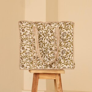 May include: A square tote bag with a floral pattern in olive green and white, set on a small wooden stool. The bag has matching handles and a stitched border. The background is a solid beige color.