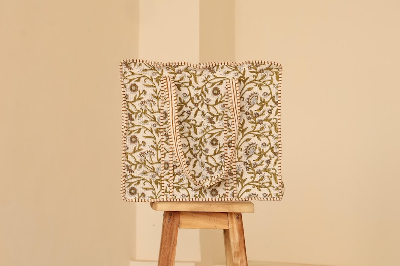 May include: A square tote bag with a floral pattern in olive green and white, set on a small wooden stool. The bag has matching handles and a stitched border. The background is a solid beige color.