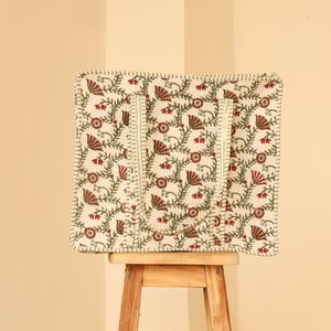 May include: A square tote bag with a floral pattern in red and green on an off-white background. The bag has two handles and a stitched border. The bag is sitting on a small wooden stool.