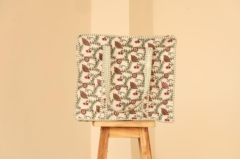 May include: A square tote bag with a floral pattern in red and green on an off-white background. The bag has two handles and a stitched border. The bag is sitting on a small wooden stool.