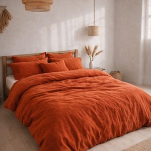 Puede incluir: Una habitación con una cama vestida con una funda nórdica y almohadas de lino de color naranja quemado. La habitación tiene una combinación de colores neutros, con pantallas de lámparas tejidas suspendidas del techo. Una alfombra tejida está al pie de la cama y un jarrón con flores secas está en la mesita de noche.