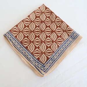 May include: A square fabric handkerchief featuring a repeating geometric pattern of rust-coloured fan shapes on a light beige background. The border consists of a blue and white floral-inspired motif that frames the central design on two sides.