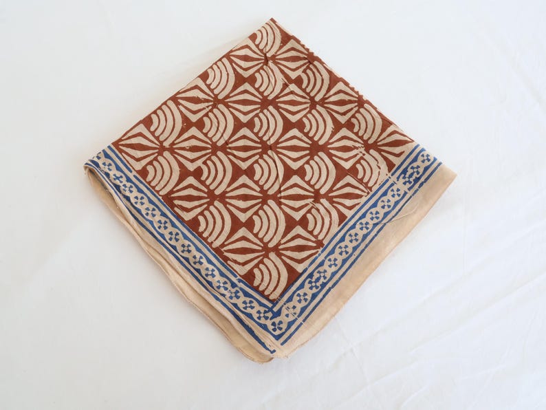 May include: A square fabric handkerchief featuring a repeating geometric pattern of rust-coloured fan shapes on a light beige background. The border consists of a blue and white floral-inspired motif that frames the central design on two sides.