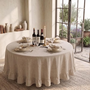 May include: A round beige tablecloth with a ruffled edge draped over a dining table. The table is set with white plates, bowls, silverware, wine glasses, three wine bottles, and a single tall white candle in a gold-toned candle holder.