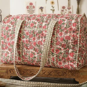 Peut inclure: Un sac de voyage à motifs floraux avec une bandoulière. Le sac présente un motif répété de fleurs rouges et de feuilles vertes sur un fond crème. Les sangles sont crème avec une bordure verte. Le sac est posé sur une table en bois.