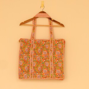 May include: An orange tote bag with a floral pattern of green leaves and white and pink flowers. The bag has matching straps and trim. It is hanging on a wooden hanger against a light orange background.