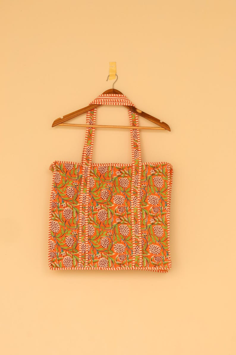 May include: An orange tote bag with a floral pattern of green leaves and white and pink flowers. The bag has matching straps and trim. It is hanging on a wooden hanger against a light orange background.
