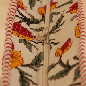 May include: Close-up of a white fabric with a floral pattern in red, yellow, and green. A silver zipper runs down the center. The edge is trimmed with red and white striped fabric.
