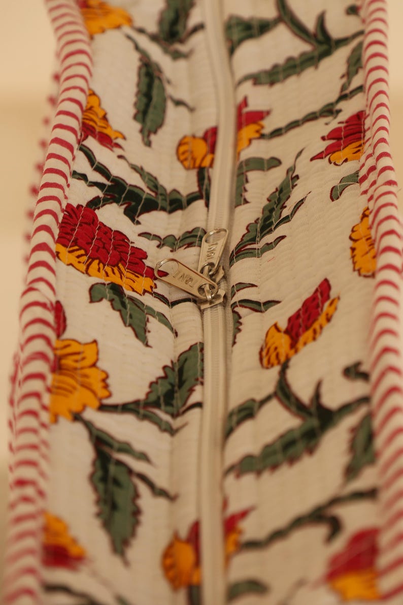 May include: Close-up of a white fabric with a floral pattern in red, yellow, and green. A silver zipper runs down the center. The edge is trimmed with red and white striped fabric.