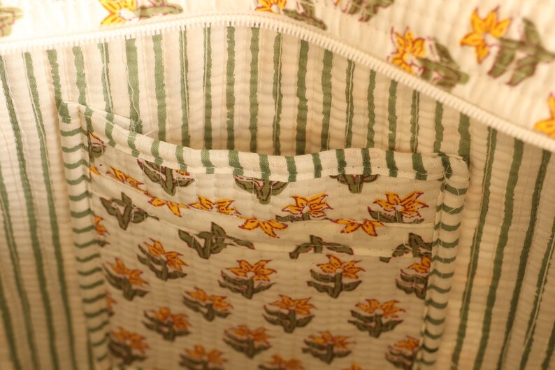May include: Interior view of a tote bag with a floral pocket. The bag's lining features vertical green stripes on a cream background. The pocket is decorated with a repeating pattern of yellow flowers and green leaves.
