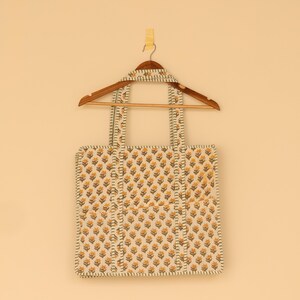 May include: A square tote bag with a floral pattern in yellow and brown on an off-white background. The bag has two handles and is hanging on a wooden hanger. The bag's edges and handles are trimmed with a striped pattern.