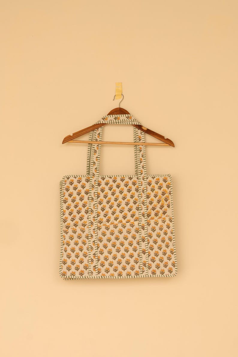 May include: A square tote bag with a floral pattern in yellow and brown on an off-white background. The bag has two handles and is hanging on a wooden hanger. The bag's edges and handles are trimmed with a striped pattern.