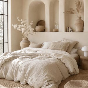 May include: A neutral-toned bedroom with a large bed covered in a linen duvet and pillows. The room features built-in wall niches with decorative vases and a large window. A woven rug and a small side table complete the space.