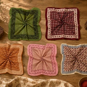 May include: Five square fabric napkins featuring block-printed patterns in olive green, deep red, burnt orange, soft pink, and cream with blue. Each napkin displays a distinct geometric or floral border and centre design, arranged on a wooden surface.