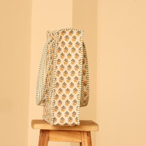 May include: A cream-colored tote bag with a repeating floral pattern in yellow and green. The bag has matching straps and is sitting on a small wooden stool. The bag's design is visible on the front and side.