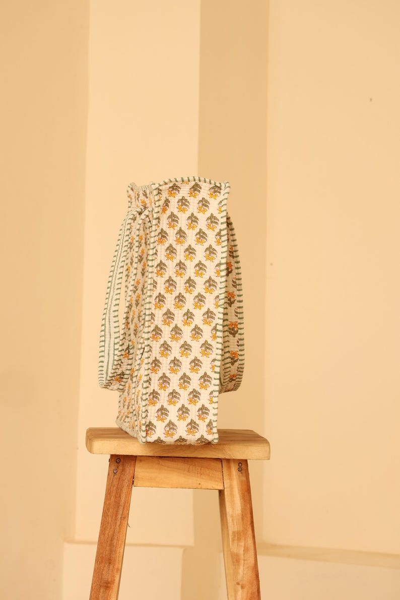 May include: A cream-colored tote bag with a repeating floral pattern in yellow and green. The bag has matching straps and is sitting on a small wooden stool. The bag's design is visible on the front and side.