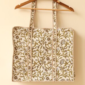 May include: Cream-colored tote bag with a floral pattern in olive green and brown, hanging on a wooden hanger. The bag features matching straps and trim, and is made of quilted fabric. The design includes stylized leaves and flowers.