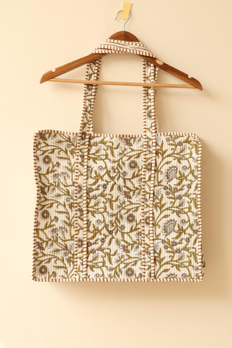 May include: Cream-colored tote bag with a floral pattern in olive green and brown, hanging on a wooden hanger. The bag features matching straps and trim, and is made of quilted fabric. The design includes stylized leaves and flowers.