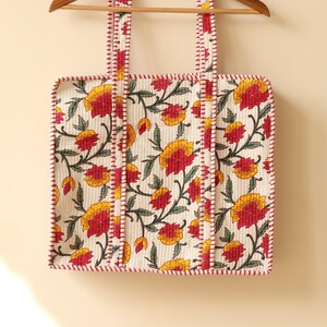 May include: Cream-colored tote bag with a floral pattern of yellow and red flowers and green leaves. The bag has red trim and straps, and is hanging on a wooden hanger.