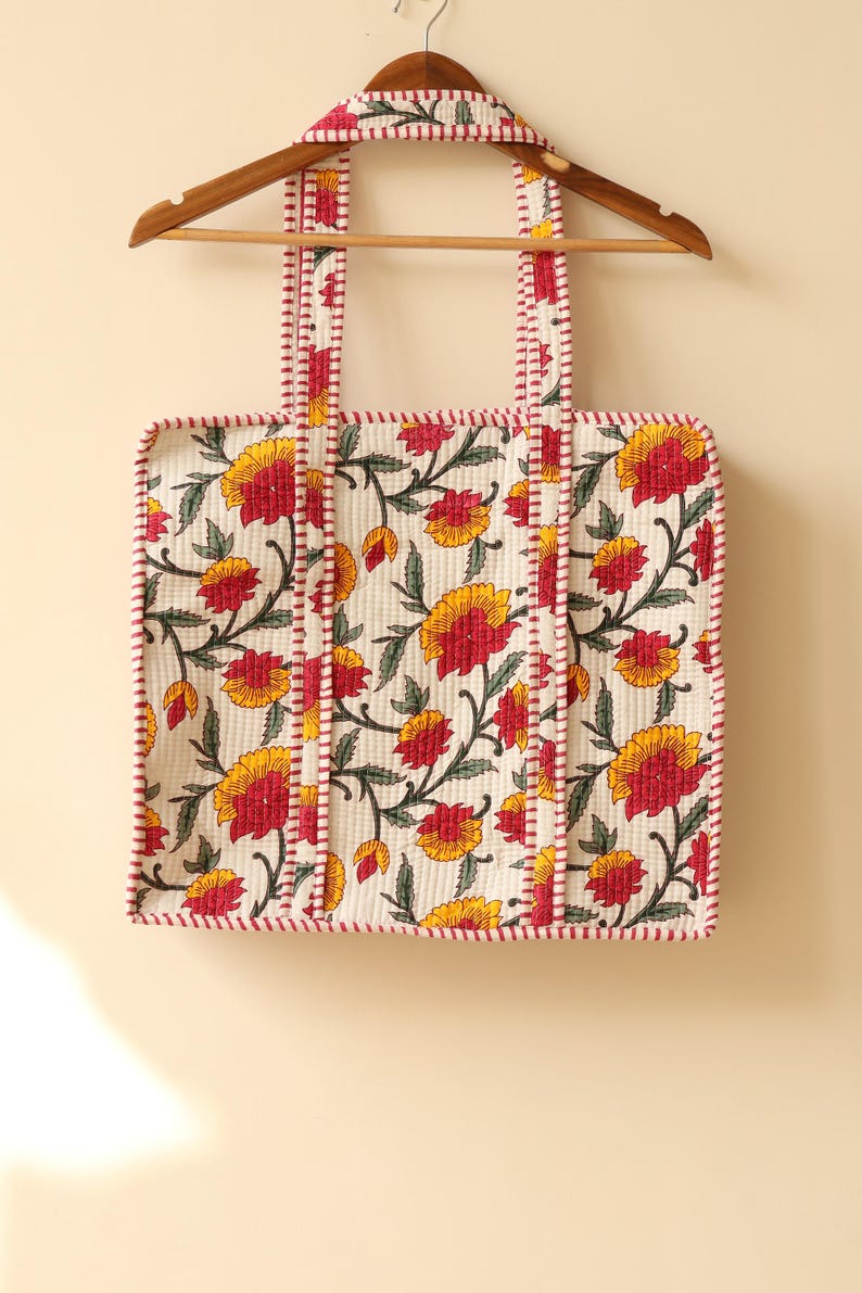 May include: Cream-colored tote bag with a floral pattern of yellow and red flowers and green leaves. The bag has red trim and straps, and is hanging on a wooden hanger.
