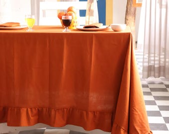 Puede incluir: Un mantel naranja con borde con volantes cubre una mesa puesta para una comida. La mesa está puesta con platos, vasos y un jarrón con flores blancas. El suelo tiene un patrón de cuadros blancos y negros.