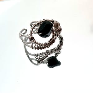 Könnte beinhalten: Ein handgefertigter, drahtgewickelter Ring mit zwei schwarzen Edelsteinen. Der Ring hat ein silberfarbenes Drahtdesign, wobei die Edelsteine in der Mitte des Rings eingesetzt sind. Der Ring wird auf einer reflektierenden Oberfläche präsentiert.