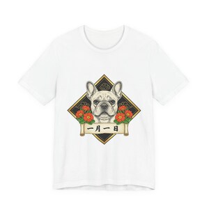 以下が含まれることがあります： 白いTシャツ。金色のひし形のフレームの中にフレンチブルドッグの顔のイラストが描かれています。赤い花と日本語のテキストが入ったバナーが犬を取り囲んでいます。デザインはシャツの中央に配置されています。
