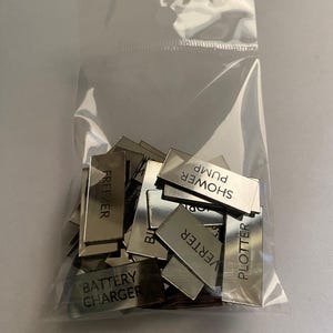 May include: A clear plastic bag filled with small, rectangular, silver-colored metal tags. Each tag has a different word printed on it, including "FREEZER", "SHOWER PUMP", and "BATTERY CHARGER".