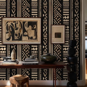 May include: An interior scene featuring a dark wooden table with books, a vase, and a small wooden stool. The wall is adorned with black and cream-colored patterned wallpaper and framed artwork. A tall, black sculpture stands to the right.