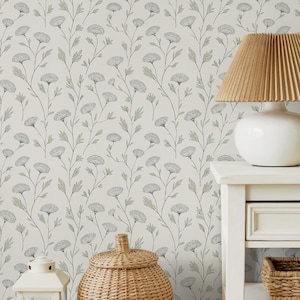 May include: A cream-colored floral wallpaper with a repeating pattern of stylized gray flowers and green leaves. A white nightstand holds a lamp with a tan pleated shade, a woven basket, and a white lantern.