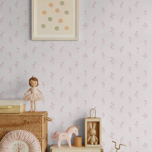 Puede incluir: Una escena de habitación infantil con una muñeca bailarina, un unicornio rosa y una bicicleta de juguete. La habitación presenta papel pintado floral y cajas de almacenamiento decorativas. Una obra de arte enmarcada con círculos coloridos cuelga de la pared. La paleta de colores es rosa suave y beige.