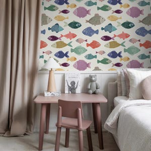 Könnte beinhalten: Kinderzimmer mit rosa Schreibtisch und Stuhl, Bett und Tapete mit bunten Fischmustern. Eine Holzlampe und ein grauer Teddybär stehen auf dem Schreibtisch. Der Raum hat beige Vorhänge und einen hellen Teppich.