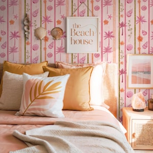 Op de afbeelding: Een slaapkamer met een strandthema. Het roze gestreepte behang heeft palmbomen, schelpen en de tekst "The Beach House". Het bed heeft oranje kussens en een wit kussen met een bladmotief. Een ingelijste strandscène hangt aan de muur.