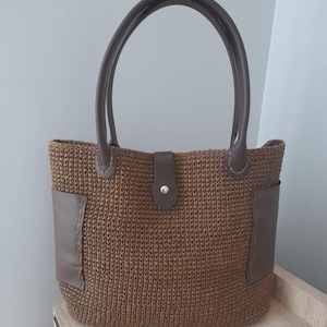 May include: A brown crocheted tote bag with brown leather accents. The bag features a top handle, a front flap with a button closure, and two side pockets. The bag is made of a woven material.