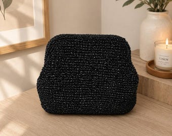 Op de afbeelding: Een zwarte clutch met een getextureerd, geweven oppervlak en een gestructureerd, afgerond rechthoekig frame. De tas heeft een consistent, dicht breipatroon over de gehele buitenkant, wat zorgt voor een uniforme uitstraling met een subtiele glans.